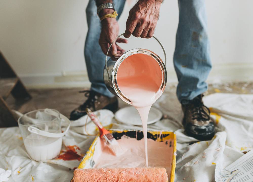 An image shows only lower part of human. A male painter is pouring a pink paint from the bucket to the paint tray. There is a small brush inside